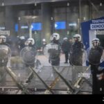 Riot police stand firm in front of a modern building, symbolizing the tense standoff between protesters and authorities amid growing European farmer protests.